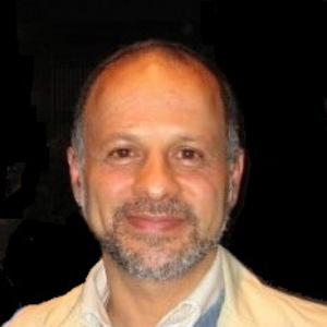 A middle-aged man with a beard and short hair smiles at the camera. He is wearing a light-colored jacket over a collared shirt, with a dark background behind him.