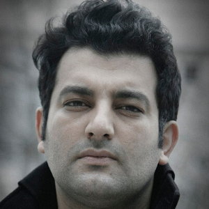 A man with short, curly black hair and a serious expression looks directly at the camera, wearing a dark jacket. The background is blurred.