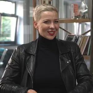 A woman with short blonde hair, wearing a black turtleneck and black leather jacket, smiles while sitting indoors with shelves of books in the background.