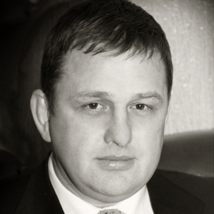 A man in a suit and tie looks at the camera with a neutral expression. The image is black and white and closely cropped to show his face and upper shoulders.
