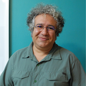 A smiling man with curly gray hair and glasses stands against a teal wall, wearing a green button-up shirt.