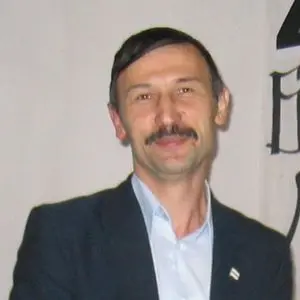 A man with short dark hair and a mustache, wearing a dark suit jacket and light-colored shirt, is standing and smiling slightly in front of a light background with partial black text visible on the side.