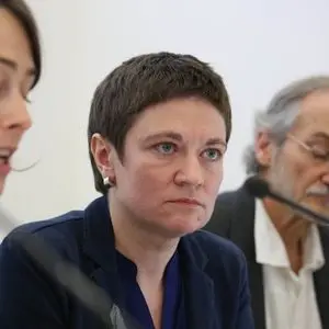 Three people sit at a table with microphones; the person in the center, wearing a navy jacket, looks serious, while the other two are partially visible and appear to be speaking or listening.