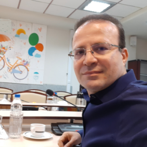 A man wearing glasses and a dark blue shirt sits at a table with a cup, glass, and bottle of water. Behind him is a colorful wall mural featuring a bicycle, balloons, and an umbrella.
