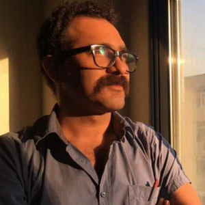 A man with glasses and a thick mustache stands by a window, looking outside thoughtfully. He wears a blue shirt and is illuminated by warm sunlight streaming in.