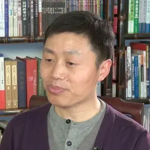 A man with short dark hair, wearing a purple cardigan over a gray shirt, sits in front of a bookshelf filled with various books.