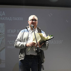 A man wearing glasses stands on stage holding a bouquet of flowers and speaking into a microphone. A projection with text and images is displayed on the screen behind him.