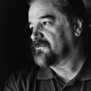 Black and white portrait of a middle-aged man with a beard and mustache, wearing a collared shirt, looking thoughtfully to the side in soft lighting.