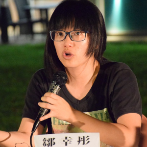 A person with straight black hair and glasses speaks into a microphone while gesturing with their hand. They are seated outdoors, wearing a dark T-shirt, with a name placard in front of them.