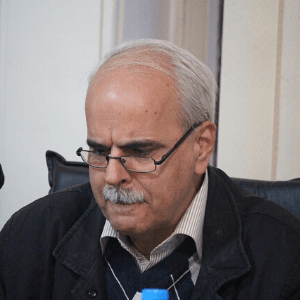 An older man with gray hair, glasses, and a mustache is sitting and looking down pensively. He is wearing a dark jacket over a striped shirt and sweater, and is indoors near a light-colored wall.