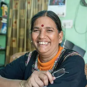 A woman with long dark hair, wearing a red bindi and black patterned top, smiles while holding glasses in her hand. She is seated indoors with shelves and a curtain in the background.