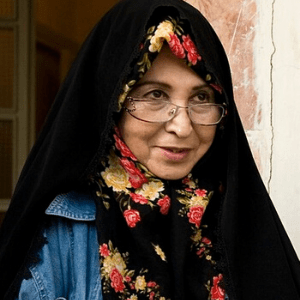 A woman wearing glasses, a black headscarf with red and yellow floral patterns, and a denim jacket stands near a light-colored wall, smiling slightly.