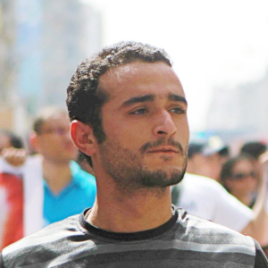 A young man with short curly hair and a trimmed beard looks ahead seriously while standing outdoors among a crowd of people. The background is bright and slightly blurred.