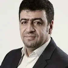 A man with short dark hair and a trimmed beard, wearing a dark suit jacket over a light-colored collared shirt, posing against a plain light background.