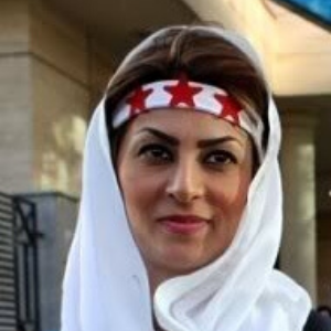 A woman wearing a white headscarf and a headband with red stars stands outdoors, smiling slightly.