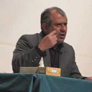 A man in a dark suit sits at a table covered with a green cloth, gesturing with his right hand. A tissue box is on the table in front of him. The background is plain and light-colored.