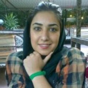 A woman with dark hair, wearing a black headscarf and a plaid shirt, sits indoors with her hand resting under her chin, smiling at the camera. Green plants and wooden furniture are in the background.