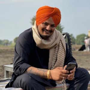 A man in a bright orange turban and black traditional attire sits outdoors, smiling and holding a smartphone. He has a light scarf draped over his shoulder and a gold bracelet on his wrist. The background is a rural setting.