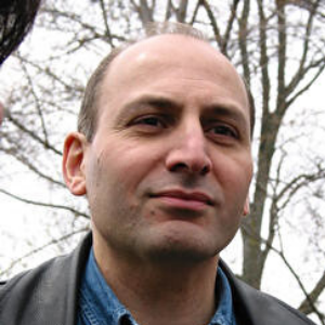 A man with short hair wearing a blue shirt and a dark jacket is outdoors, with bare trees visible in the background on a cloudy day.