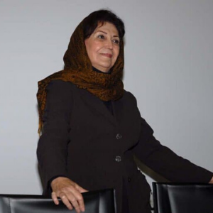 A woman wearing a dark suit and a brown patterned headscarf stands and smiles, resting her hands on the back of two black chairs against a plain background.