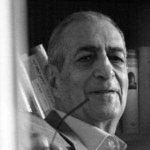 Black and white photo of an older man smiling slightly, holding a pair of eyeglasses. He is posed in front of a bookshelf filled with books.