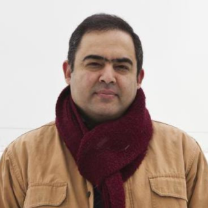 A man with short dark hair and a maroon scarf is wearing a tan jacket, standing against a plain white background, looking at the camera with a neutral expression.