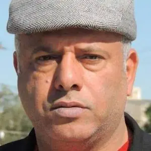 A man wearing a light gray flat cap and a black jacket looks directly at the camera with a serious expression. The background is outdoors and slightly blurred.