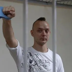 A man with a mohawk hairstyle stands behind metal bars, raising his right fist and holding a blue cloth. He wears a white graphic t-shirt and has a serious expression on his face.
