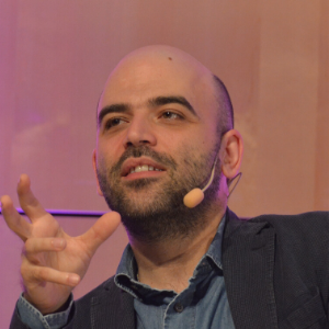 A man with a beard and shaved head speaks while gesturing with one hand. He is wearing a headset microphone, a blue shirt, and a dark blazer. The background is softly lit in warm tones.