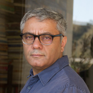 A middle-aged man with short gray hair and glasses, wearing a blue collared shirt, looks seriously at the camera. There are blurred shelves of books and a window in the background.