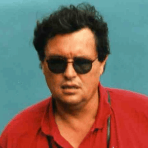 A man with dark hair wearing black sunglasses and a red shirt stands in front of a teal background. He has a neutral expression and a pair of sunglasses hanging from his neck.