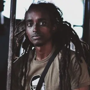A person with long dreadlocks looks directly at the camera, wearing a brown t-shirt and a beaded necklace. The background is dimly lit and out of focus.