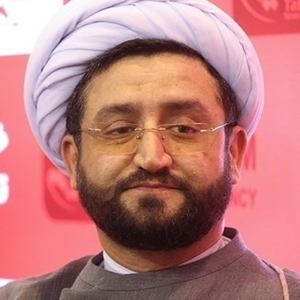 A man with a dark beard and glasses wears a white turban and gray robe, standing in front of a red backdrop with white text and logos.