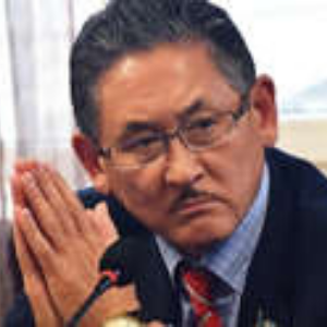 A middle-aged man wearing glasses and a suit sits at a table with his hands clasped, looking serious and focused. A microphone is visible in front of him.