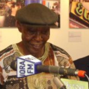 An older man wearing a patterned shirt and flat cap smiles while speaking at a microphone labeled NORA FM, with colorful artwork visible in the background.