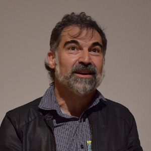 A middle-aged man with dark hair and a gray beard, wearing a patterned shirt and a black leather jacket, is looking slightly upward and smiling, standing against a plain light-colored background.
