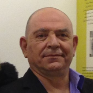 A middle-aged man with a shaved head and a slight smile is wearing a black jacket over a light purple shirt. He is standing indoors in front of a wall with text and images.