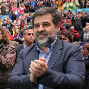 A man with short dark hair and a beard, wearing a suit, is standing and clapping in a crowded indoor setting with many people in the background.