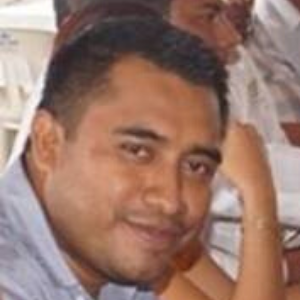A man with short dark hair and a trimmed beard is smiling slightly, looking toward the camera. He is wearing a light-colored collared shirt. Other people and white chairs are blurred in the background.