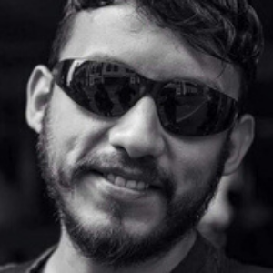 A man with a beard and mustache wearing dark sunglasses smiles slightly. The image is in black and white.