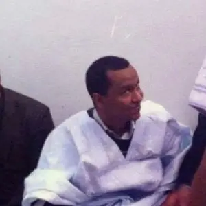 A man wearing a light blue traditional robe sits indoors, looking to his right with a slight smile. The background is plain and light-colored.