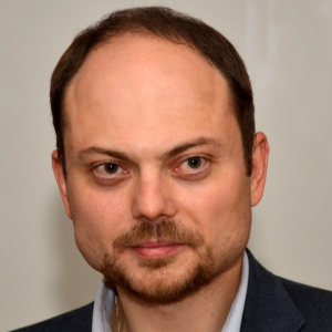 A man with light skin, short brown hair, and a trimmed beard and mustache, wearing a dark jacket and light shirt, looks directly at the camera against a plain background.