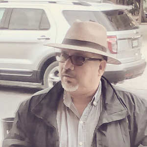 A man with glasses and a goatee wears a brown fedora and a dark jacket over a striped shirt, standing outdoors in front of a parked SUV.