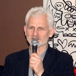 A man with short white hair and a beard speaks into a microphone. He is wearing a dark blazer over a striped shirt, standing in front of an abstract black and white artwork featuring faces and hearts.