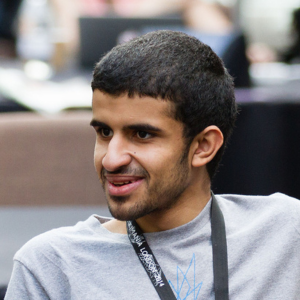 A young man with short dark hair and a trimmed beard is smiling while sitting indoors, wearing a grey T-shirt and a black lanyard around his neck. The background is blurred with people and tables.