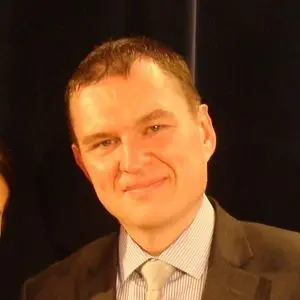 A man with short brown hair, wearing a dark suit, light blue dress shirt, and light-colored tie, smiling in front of a dark background.