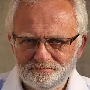 An older man with white hair and a full white beard is looking directly at the camera. He is wearing glasses and a white collared shirt. The background is out of focus.