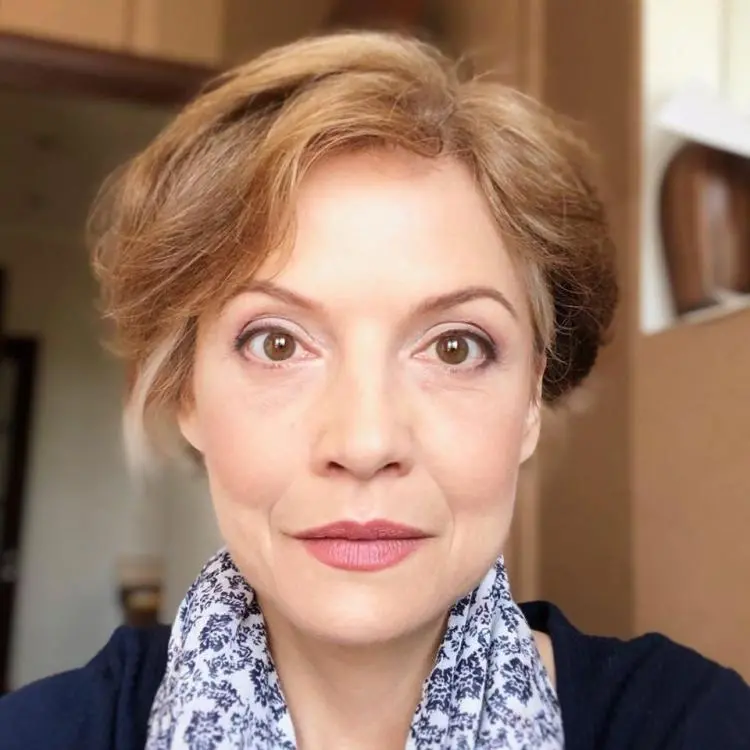 A woman with short, light brown hair and brown eyes looks directly at the camera. She is wearing light makeup and a blue and white patterned scarf, with a soft background indoors.