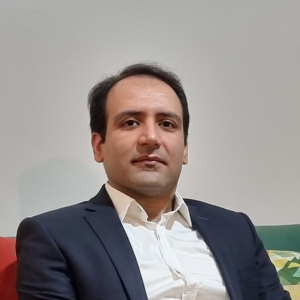 A man with short dark hair, wearing a white shirt and dark suit jacket, sits on a colorful sofa against a plain light-colored wall, looking directly at the camera with a neutral expression.