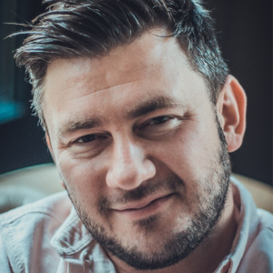 A man with short dark hair and a beard smiles softly at the camera. He is wearing a light-colored collared shirt and is seated indoors with a blurred dark background.